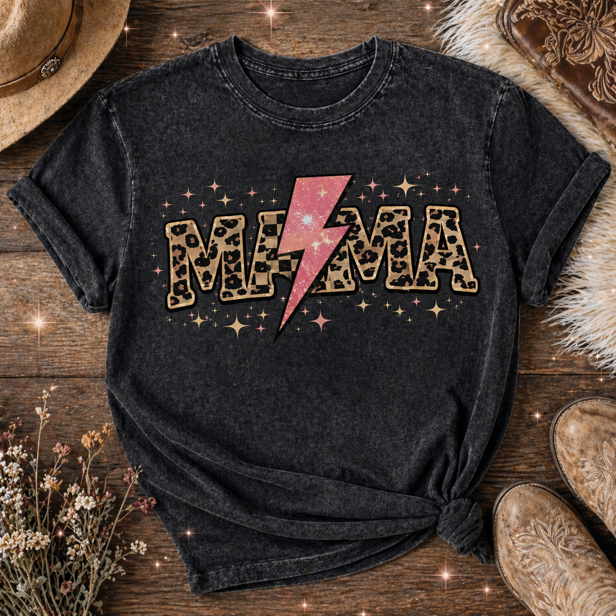 Dark charcoal mama graphic t-shirt styled in a rustic flat lay with cowboy boots, hat, dried flowers, and wooden background featuring leopard print MAMA lettering with a pink lightning bolt and sparkles.