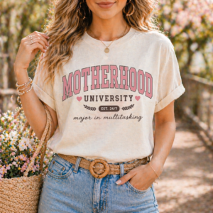 Cream graphic t-shirt featuring the phrase Motherhood University in pink collegiate lettering with hearts, established 24/7 text, and major in multitasking script for a trendy mom inspired design.